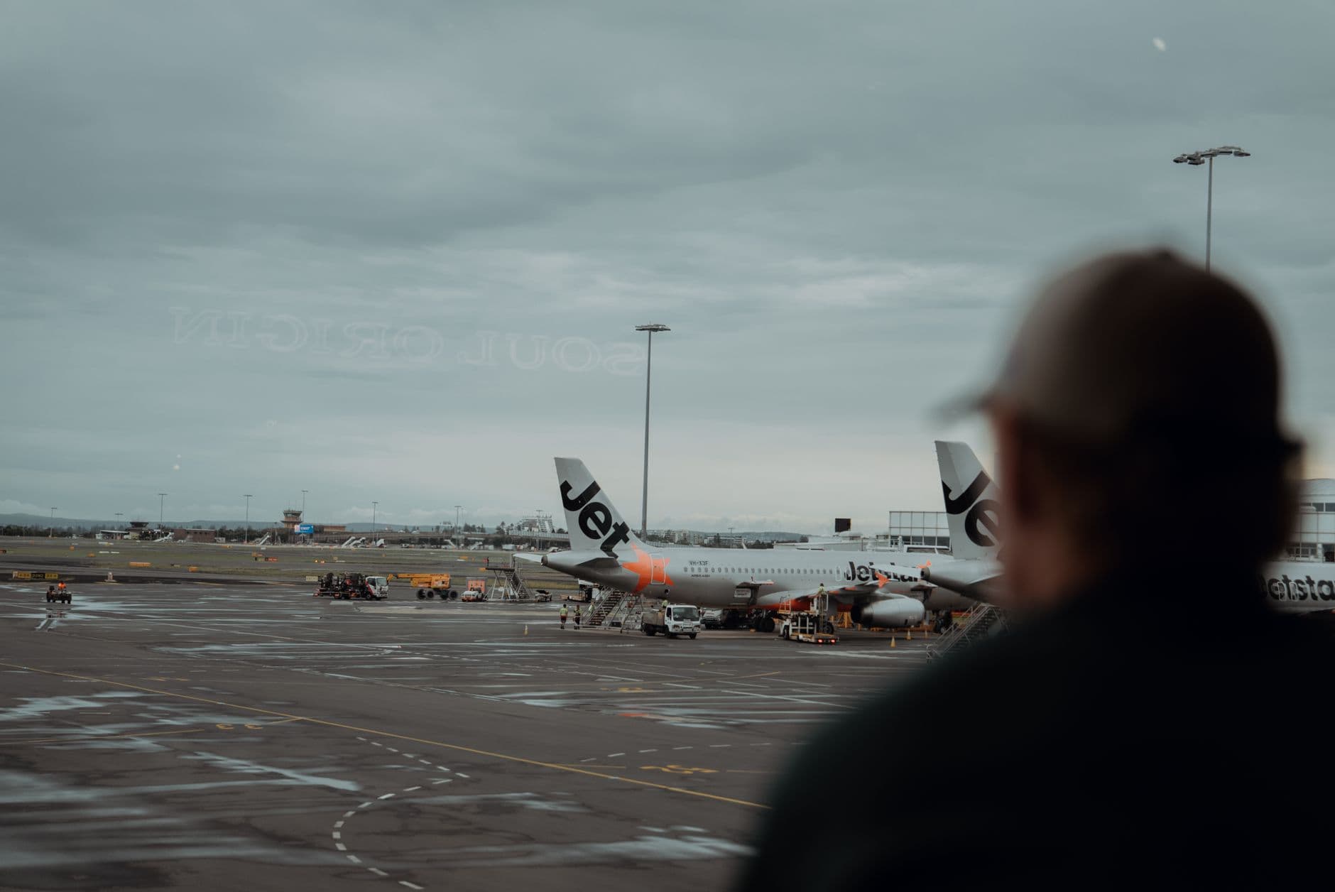 View of Jetstar airplanes parked at Sydney Airport with overcast skies and a passenger silhouette.